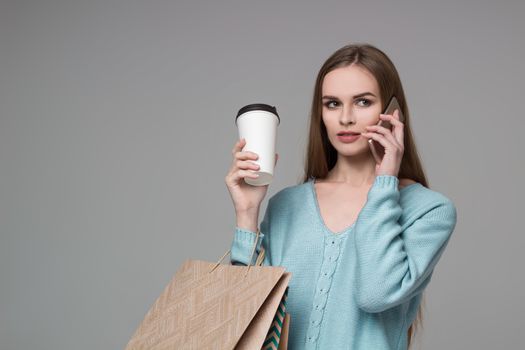 Young model long-haired blond girl in light blue aquamarine jamper holds shopping paper bags and  plastic coffee glass drinking and talking by moble smartphone