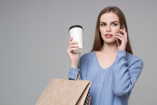 Young model long-haired blond girl in light blue jamper holds shopping paper bags and  plastic coffee glass drinking and talking by moble smartphone