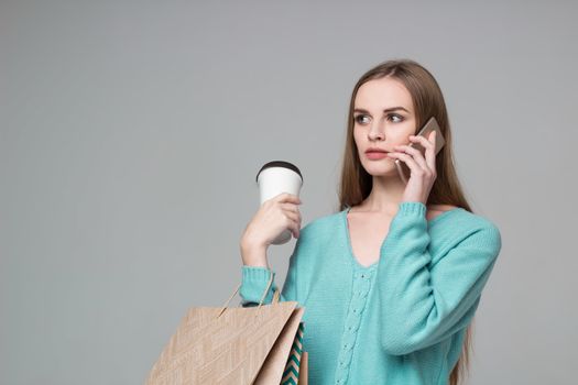 Young model long-haired blond girl in light blue aquamarine jamper holds shopping paper bags and  plastic coffee glass drinking and talking by moble smartphone