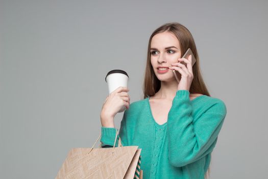 Young model long-haired blond girl in light blue aquamarine green jamper holds shopping paper bags and  plastic coffee glass drinking and talking by moble smartphone
