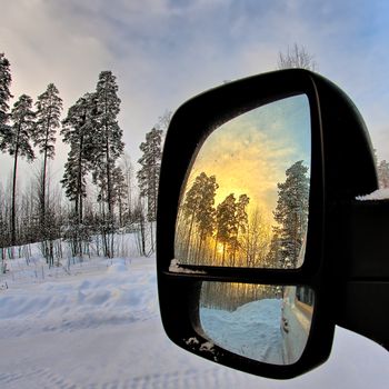 Winter forest landscape on background and beautiful yellow sunrise in rear view mirror.