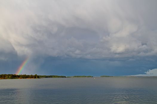 Thunderstorm approaching. Dark clouds hanging over silent water surface, covering most of the colorful rainbow. Stormy afternoon in summertime.