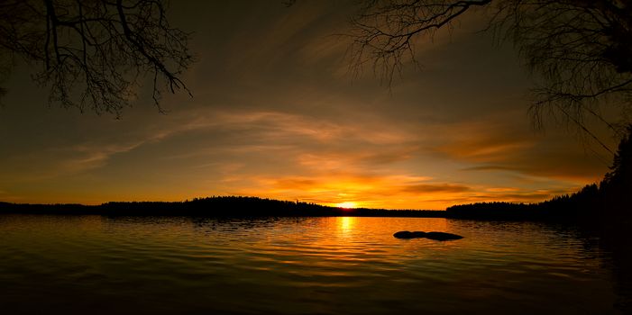 A beautiful wide angle photo of the sun setting behind the horizon of the lake. Strong amber color summer sunset by the water.