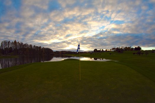 Dark golf course shortly after the sunset. Sun still glowing on clouds, lonely flag still standing at the golf course.