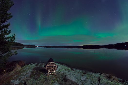 Sitting in the chair next to the water at night. Bright green aurora borealis in the sky.