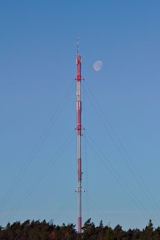 High red and white cellular tower at daytime with moon shining next to the top of the tower.