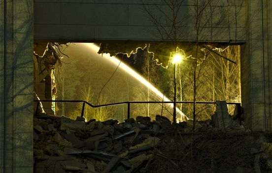 A view through collapsed building with only wall still standing. Water from broken water pipe creating a fountain lighted by street lights.
