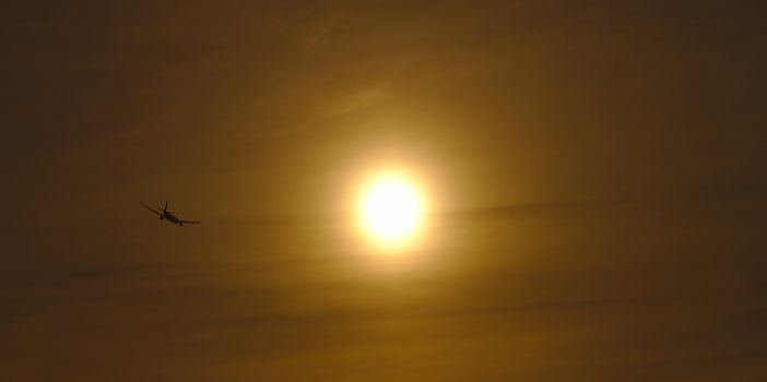 Plane flying away into the evening sun. Dark silhouette of the aircraft visible on the left side of large foggy sun.