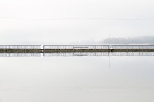 Clear reflection of a walking bridge on the surface of silent water early in the morning. Background covered in fog.