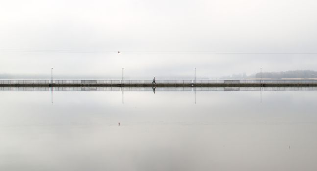 Lonely walker on a long empty bridge early in the morning. Grey and foggy weather in the city.