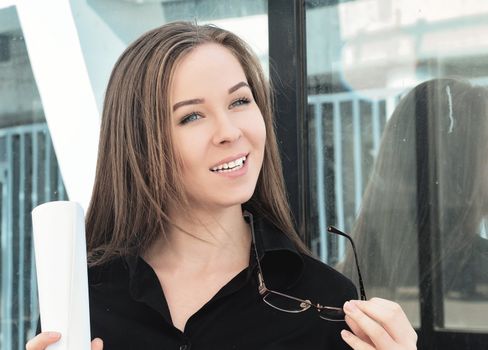 Portrait of a beautiful young engineer girl who holds a sheet of paper and glasses, happy to complete the project, standing near the building in the open, close-up