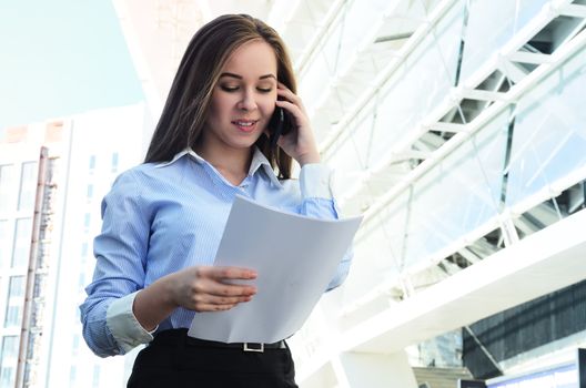 Young beautiful and successful business lady keeps sheets of paper talking on the phone, being near the business center