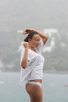 Attractive young female in while blouse and panties embracing herself and keeping eyes closed while standing near water on windy day
