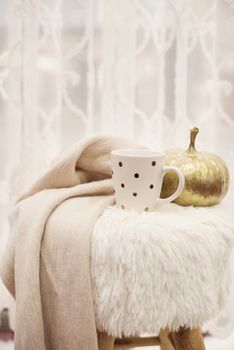 Hot chocolate, a cup of cappuccino on a fur chair in front of a large window with a white sheers curtain. Warm scarf and golden pumpkin around. Cozy winter evenings
