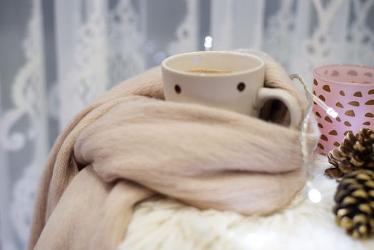Hot chocolate, a cup of cappuccino on a fur chair in front of a large window with a white sheers curtain. Warm scarf, cones and lights around. Cozy winter evenings