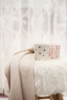 Hot chocolate, a cup of cappuccino on a fur chair in front of a large window with a white sheers curtain. Warm scarf and lights around. Cozy winter evenings