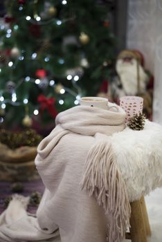 Christmas time. Hot chocolate, a cup of cappuccino on a fur chair in front of a large Christmas tree with balls and lights. Warm scarf, cones around