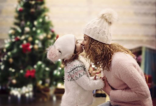 Happy Christmas and Happy Holidays concept. Cheerful mom and her cute daughter having fun in cozy decorated living room near the Christmas tree. Mother is kissing, hugging the little girl.