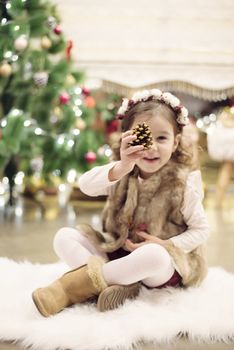 Cute little girl celebrating Christmas at home. She sits on a chair and holds a spruce cone in her hands. Positive emotions. Christmas tree in background. Happy childhood.