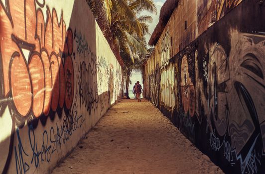 The very narrow alley covered with graffiti leading to the beach of Playa del Carmen in Mexico, with two people walking towards the exit.