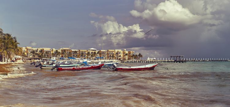 Several small fishing boats are moored by the sea in the beautiful beach of Playa del Carmen in Mexico with the background of the typical buildings of this place.