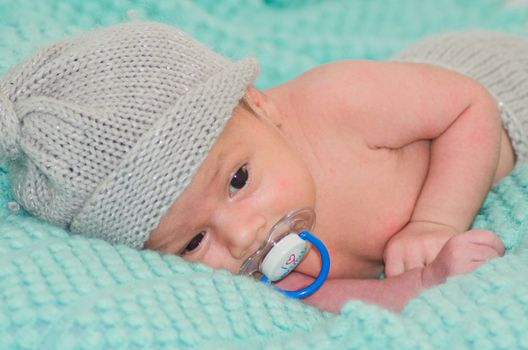 4 weeks old newborn baby boy with gray hat on green blanket with dummy in his mouth