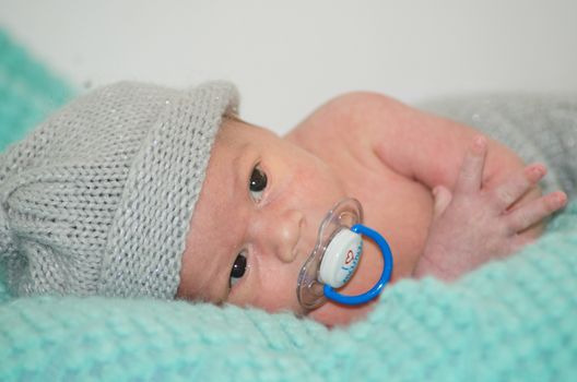 4 weeks old newborn baby boy with gray hat on green blanket with dummy in his mouth