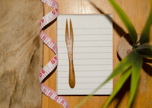notebook with wooden fork and measuring tape on wooden table, diet and healty eating concept