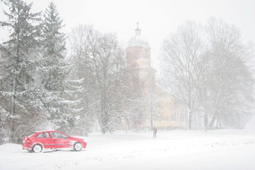Church in snowfall. Winter in Vecsaule, Latvia. Church covered in snow. Winter landscape with snow covered church, trees and car.



