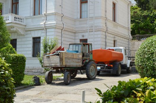 Old tractor with garbage in the back is near the watering machine, cleaning equipment for cleanliness in parks on a Sunny summer day.