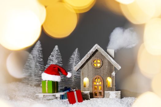 Christmas card with cozy small decorative house , gifts and Santa Claus cap on sleigh on snow over fairy tale winter fir forest background and falling snow . New Year design postcard . Copy space for text