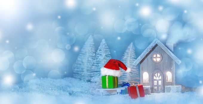 Christmas card with cozy small decorative house , gifts and Santa Claus cap on sleigh on snow over fairy tale winter fir forest background and falling snow . New Year design postcard . Copy space for text