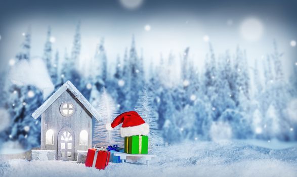Christmas card with cozy small decorative house , gifts and Santa Claus cap on sleigh on snow over fairy tale winter fir forest background and falling snow . New Year design postcard . Copy space for text