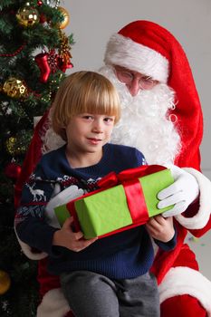 Portrait of smiling little boy sitting on santa claus knees and getting a gift