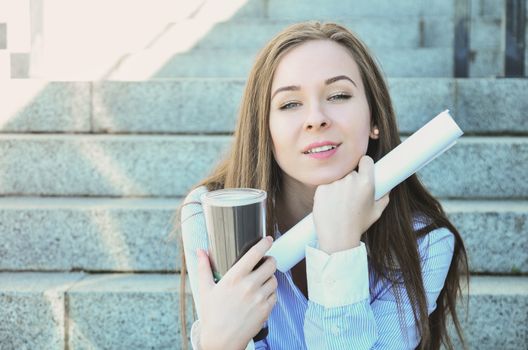 Beautiful attractive girl student, sat down on the steps during the break, with a hot thermos in her hands, holding a bundle of drawings for the project