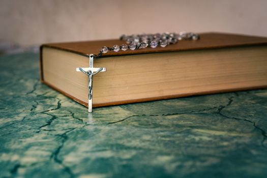 Silver rosary and cross resting on the closed book at green table,front view.religion school concept.Vintage style.