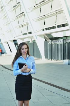 Beautiful young business woman standing and holding a notebook, wearing a black skirt and shoes, looking into the distance, vertical photo