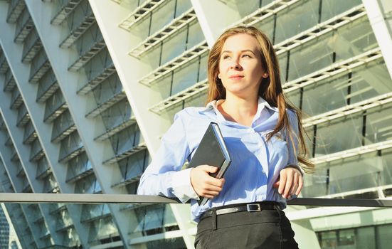 Young beautiful business lady standing in office clothes leaning on the railing, and holding a notebook and peering into the distance