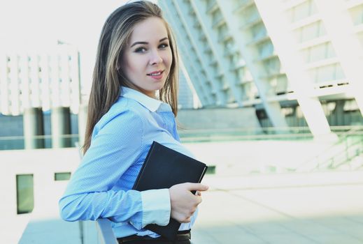 Cheerful girl student in beautiful office clothes, standing near the railing and holding a black notebook, while looking at the camera and smiling