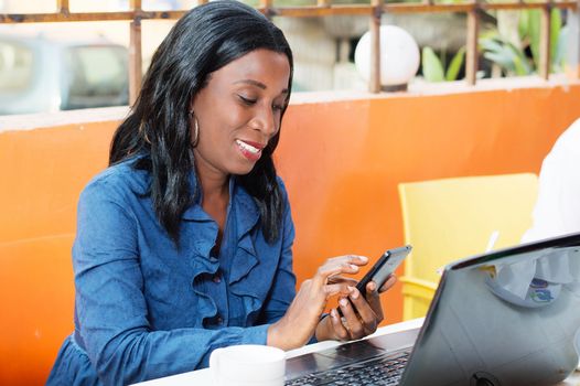 This smiling young woman sitting at a laptop and manipulates her mobile phone.
