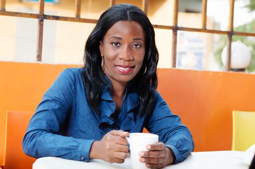 smiling young woman is sitting in the restaurant and holding a cup of coffee in hands.
