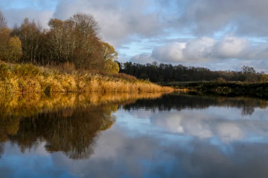 View on autumn landscape of river and trees in sunny day. Forest on river coast in autumn day. Reflection of autumn trees in water. Autumn in Latvia. Autumn landscape with colorful trees and river. 