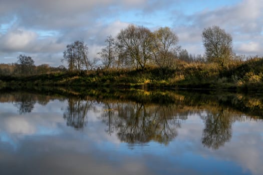 View on autumn summer of river and trees in sunny day. Forest on river coast in autumn day. Reflection of autumn trees in water. Summer in Latvia. Autumn landscape with colorful trees and river. 