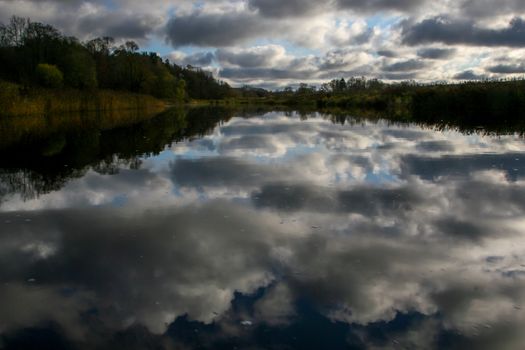 View on summer landscape of river and trees in evening. Forest on river coast in summer day. Reflection of clouds and sky in water. Summer in Latvia. Autumn landscape with colorful trees, grass and river. 