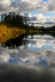 View on autumn landscape of river and trees in sunny day. Forest on river coast in autumn day. Reflection of clouds in water. Autumn in Latvia. Autumn landscape with colorful trees, yellow grass and river. 