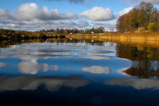 View on autumn landscape of river and trees in sunny day. Forest on river coast in autumn day. Reflection of autumn trees in water. Autumn in Latvia. Autumn landscape with colorful trees, yellow grass and river. 