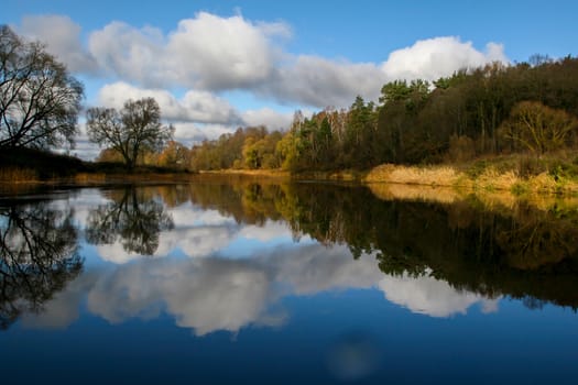 View on autumn landscape of river and trees in sunny day. Forest on river coast in autumn day. Reflection of autumn trees in water. Autumn in Latvia. Autumn landscape with colorful trees, yellow grass and river. 