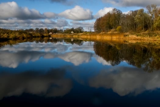 View on autumn landscape of river and trees in sunny day. Forest on river coast in autumn day. Reflection of autumn trees in water. Autumn in Latvia. Autumn landscape with colorful trees, yellow grass and river. 