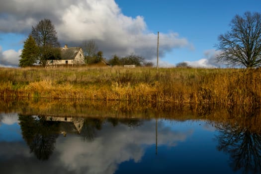 View on autumn landscape of river and trees in sunny day. Grass on river coast in autumn day. Reflection of autumn grass and house in water. Autumn in Latvia. Autumn landscape with colorful trees, yellow grass and river. 