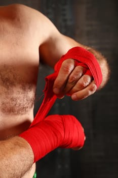 Close up man boxer wrapping red hand wraps over wrists preparing for fight, over black background with copy space, low angle view